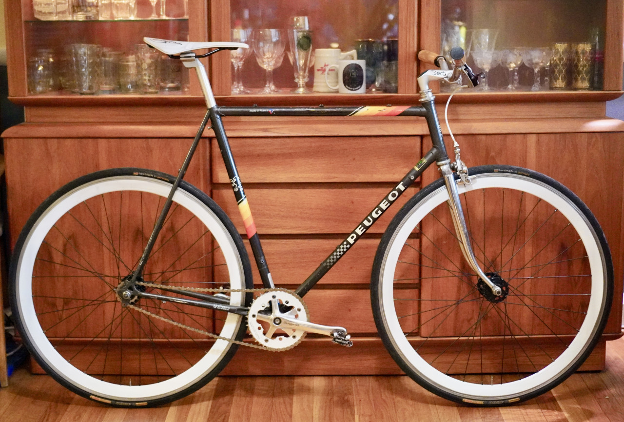 A fixed gear bike made from a vintage black Peugeot steel frame with orange/yellow accents and a polished chrome fork stands on a wooden floor in front of a mid-century Danish cabinet. The bike has a white racing saddle, a short Deda stem with riser flat bars and wooden grips fitted with one brake lever on the right side attached to a front Ultegra brake caliper. The wheels have deep white sections and are fitted with Continental GP4000 23C tires. The Origin8 chainring is white 42T fitted with Sugino RD2 "Messenger" crank arms, and is mated to a 15t cog attached to a flip-flop rear hub. 
