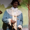 A wax statue of a black man in the garb of a 17th century aristocrat - a black napoleonic hat with am orange flower, a light blue suit with orange edging and dripping with lace - stands in front of a painted outdoor scene.