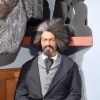 A wax statue of a  black man with a frankly wild afro picked out to extraordinary volume that has a white streak over his right eye, and a bushy mustache and goatee sits wearing a grey suit with a black tie and black overcoat. His expression is stern and serious.