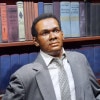 A wax statue of a black man with a square face and, true to life, a kind expression, wearing a gray suit and tie sits in front of a wall painted to look like a bookshelf.