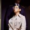 A wax statue of a black woman in a trench coat and oversized glasses is led down the stairs of a bus in handcuffs by the driver (also a statue).