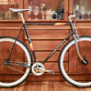 A fixed gear bike made from a vintage black Peugeot steel frame with orange/yellow accents and a polished chrome fork stands on a wooden floor in front of a mid-century Danish cabinet. The bike has a white racing saddle, a short Deda stem with riser flat bars and wooden grips fitted with one brake lever on the right side attached to a front Ultegra brake caliper. The wheels have deep white sections and are fitted with Continental GP4000 23C tires. The Origin8 chainring is white 42T fitted with Sugino RD2 "Messenger" crank arms, and is mated to a 15t cog attached to a flip-flop rear hub. 