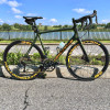 A bicycle is parked on the side of the road, leaning against a curb. It is a green and black bike with a yellow-orange logos on the downtube and wheesl. The bike is positioned near a fence and a body of water, creating a scenic backdrop.