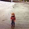 A red-headed toddler wearing a red and black striped sweater and jeans stands on a wet beach close to where the waves are meet the sand. There is a boat in the distance and twigs, rocks and debris on the sand. 