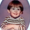 A 4 year-old boy with red hair in a Dutch boy cut wearing a striped shirt looks to the right and smiles. 