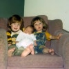 Two young children sit on a big overstuffed arm chair while an infant is propped between them. On the left a boy with a longish red hair wears a striped shirt and tries to smile. On the right a girl in overalls makes an O with her mouth and looks a little surprised. The infant in the middle has his eyes closed and seems to be asleep in a white set of pajamas.