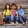 Three children sit on a set of steps. The two on the left are east asian while the boy on the left is white. The girl on the far right has long dark hair and a yellow shirt and is smiling broadly. The boy in the middle has short dark hair and a red striped shirt and is smiling but seems concerned. The boy on the right wears a blue sweatshirt, has longish red hair in a dutch boy cut and has his arm around the arm of the boy next to him as he leans into him smiling as big as he can.