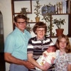 A trio of people in decreasing height from left to right stand with the woman in the middle holding a baby. They all smile at the camera. The man with glasses on the left is middle-aged and has dark hair that is highly Brylcreemed and he is sturdy and well-built. The girl on the right is a young teen with sandy blonde hair and a patterned shirt. The woman in the middle is also middle-aged and medium brown hair that is cut short with bangs. She wears glasses and is holding a very pouty-faced infant wrapped up in blankets.