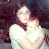 A 25-year-old woman with long dark hair and glasses who could pass for 16 holds a 4-month-old infant who is gnawing on his left fist. The woman has a far away look in her eyes and there are Christmas presents behind her. The image is heavily damaged with many speckles where the surface has been worn away.