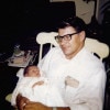 A smiling white man with dark hair in a very Byrlcreemed haircut  sits smiling in a rocking chair while holding a baby in his lap in a very casual pose.