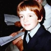 A 7-year-old white boy with red hair and many freckles wearing a navy blue suit with a light blue shirt looks smiling to the right as he sits in a church pew. A woman is sitting behind the boy and holding a bulletin.