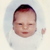 A white newborn infant with a slightly misshapen head and a little more hair than you might expect looks directly at the camera. He has a scratch on his left eyelid and is raising his left hand.