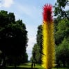 A large, colorful, and tall sculpture made of colored glass is on display in a park. The sculpture is located in the middle of a grassy field, surrounded by a fence. There are several people in the park, walking around and enjoying the outdoors. Some of them are closer to the sculpture, while others are further away. The overall atmosphere of the park is lively and inviting.