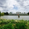 A large building with a glass roof is situated on a lake, surrounded by a lush green garden. The building appears to be a greenhouse. There are several people scattered around the area, enjoying the view and the peaceful atmosphere. The scene is further enhanced by the presence of a statue with a red blown glass flower in the center of the lake, adding to the overall beauty of the location.